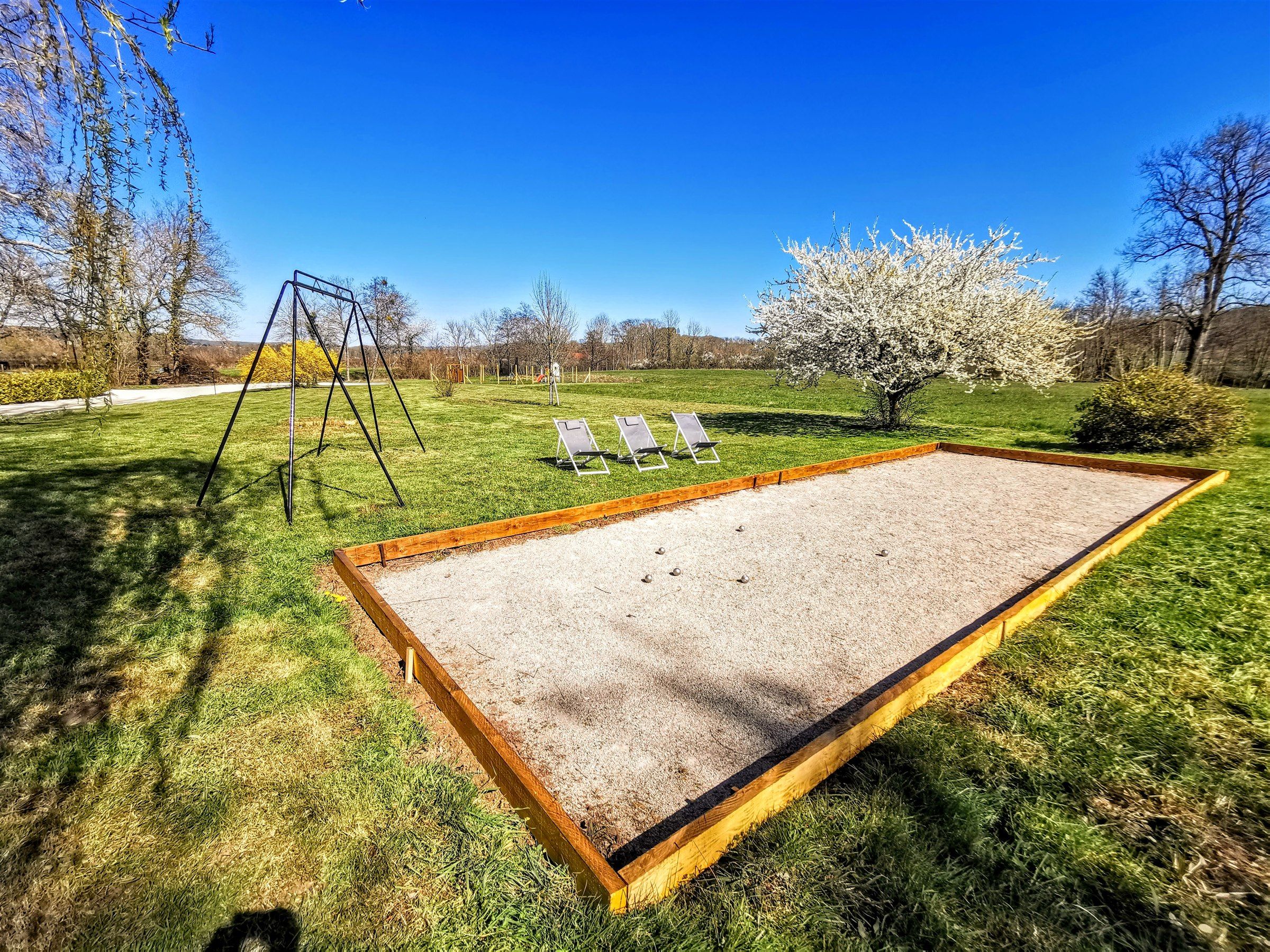 Un jardin avec une balançoire, deux chaises de jardin, un bac à sable et un arbre.
