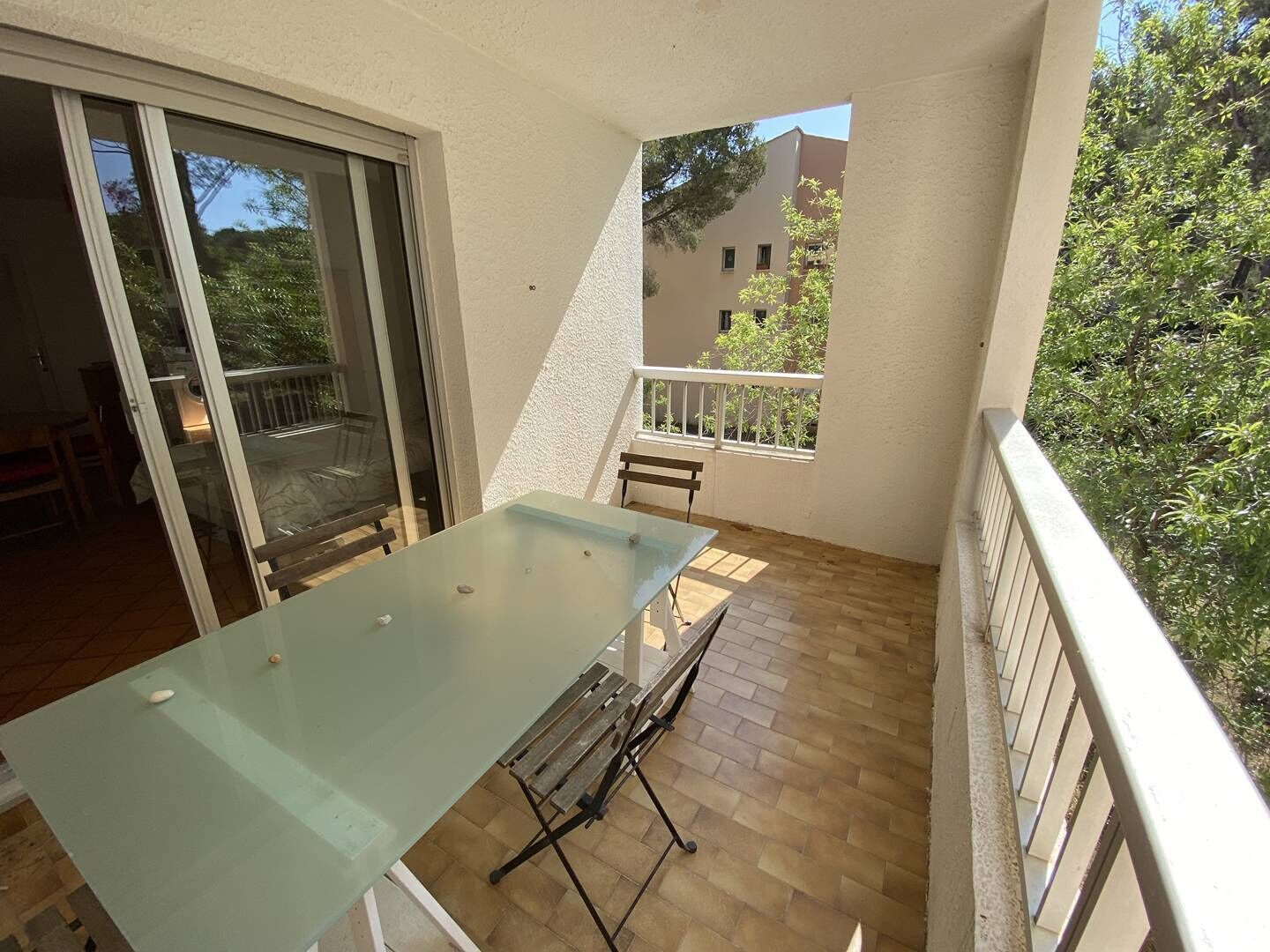Un balcon doté d’une table à plateau en verre et de bancs, d’une porte-fenêtre coulissante, et offrant une vue sur un immeuble et des arbres.