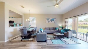 A spacious living room with a gray sofa, a dining area with a table and chairs, and a large sliding glass door leading to a balcony. - PERFECT, NEW! 6 Min to Dis, On Lake, Brand Name Beds, Big  T.Vs, Steps to Pool.  (Kissimmee)