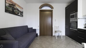 A living room with a dark sofa, a white chair, a wooden door, and a kitchen with black cabinets and a microwave. - Italianway - Andrea Ponti 1 (Milan)