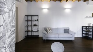 A living room with a grey sofa, a black shelving unit, and a round white ottoman. - Italianway Comfort - Alfieri 18 (Milan)
