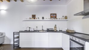 A modern kitchen with white cabinetry, a black countertop, and a built-in oven. There are shelves above the counter holding various kitchen items and a framed picture. - Italianway Comfort - Alfieri 18 (Milan)