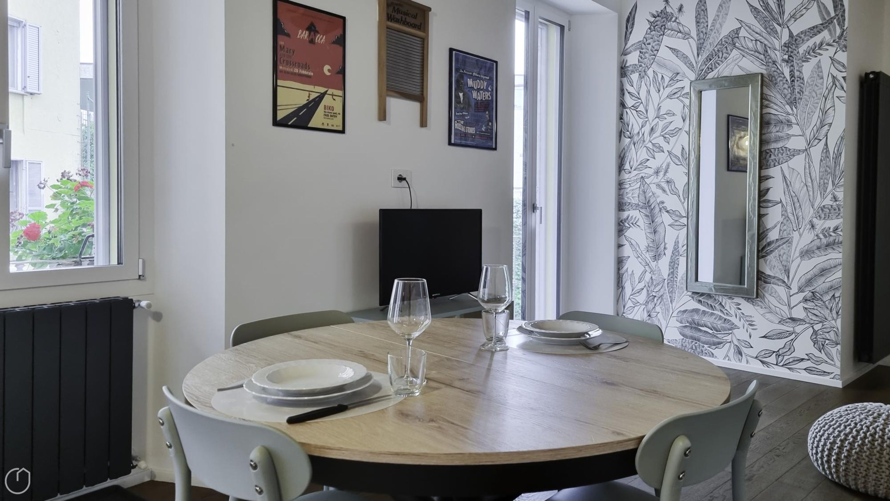 Un coin repas avec une table ronde en bois, dressée pour deux personnes, deux verres à vin et des assiettes. On y trouve deux chaises, un radiateur et un téléviseur fixé au mur.