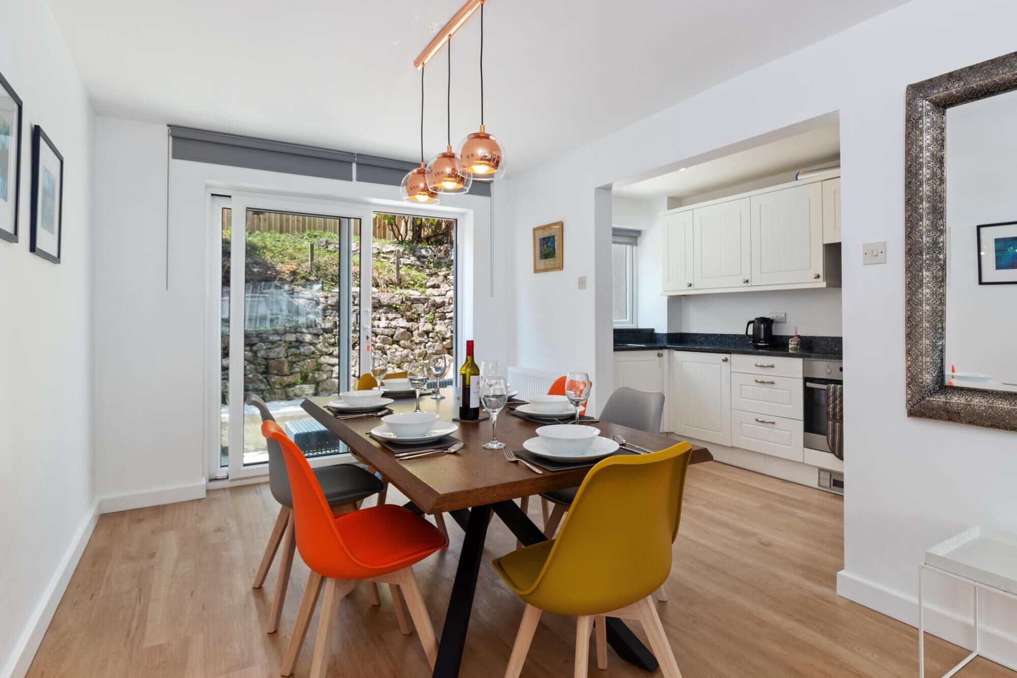 A dining area with a wooden table, orange and yellow chairs, and a view of a stone wall and greenery outside.