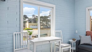 A room with a white table and chairs, a small round table with a plant, a grey sofa, and a window with a view of houses and trees.