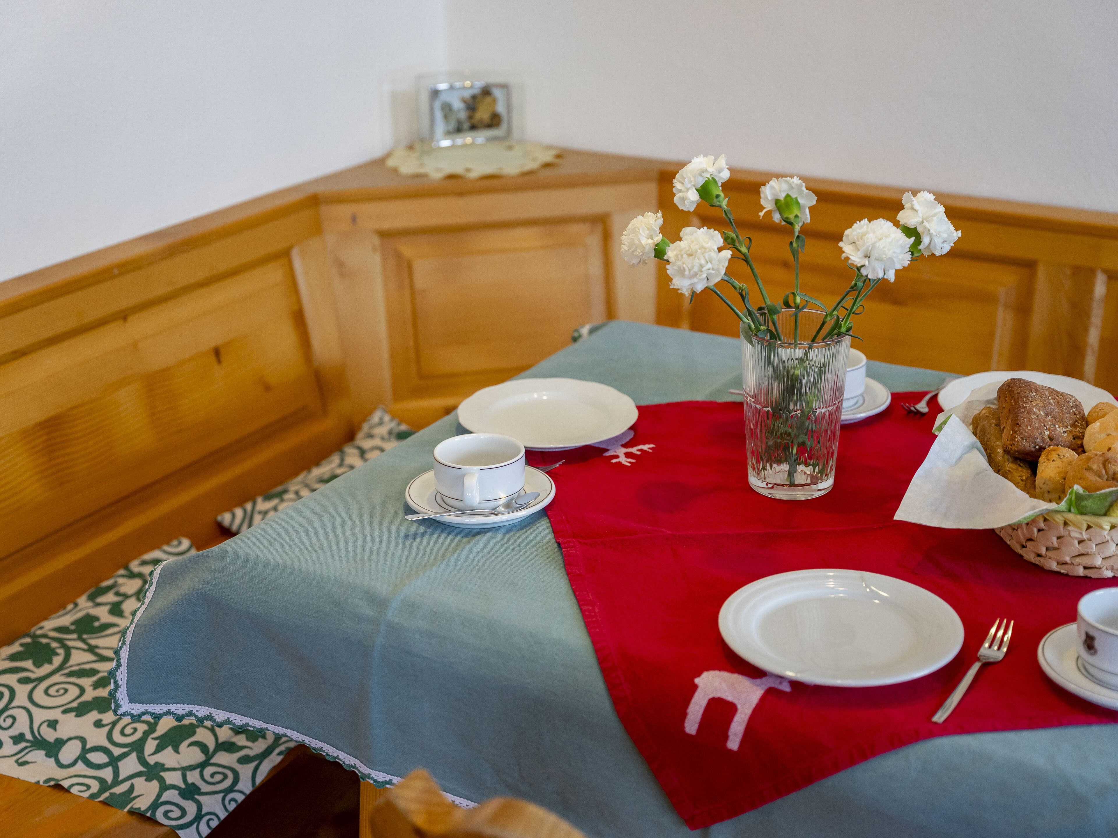 A dining table with a red placemat, white plates, a glass of water, a cup, and a basket of bread.