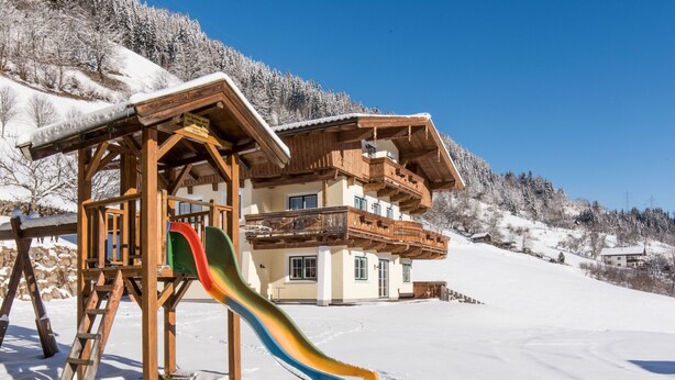 Eine Holzhütte mit einer bunten Rutsche vor einer verschneiten Berglandschaft.
