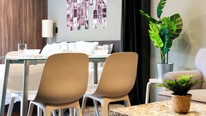 Une chambre d’hôtel moderne dotée d’un coin repas comprenant une table en verre, des chaises blanches et une grande plante en pot.