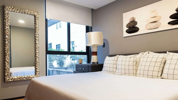 Una habitación de hotel moderna con una cama grande, una mesita de noche con lámpara, un espejo con marco decorativo y una obra de arte en la pared representando piedras apiladas.
