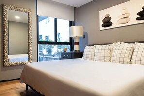 Et moderne hotelværelse med en stor seng, et natbord med lampe, et spejl med dekorativ ramme og et kunstværk bestående af stablede sten.