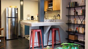 Una cocina compacta con refrigerador, microondas y pileta. Hay una pequeña isla con taburetes, una estantería y una planta en maceta.