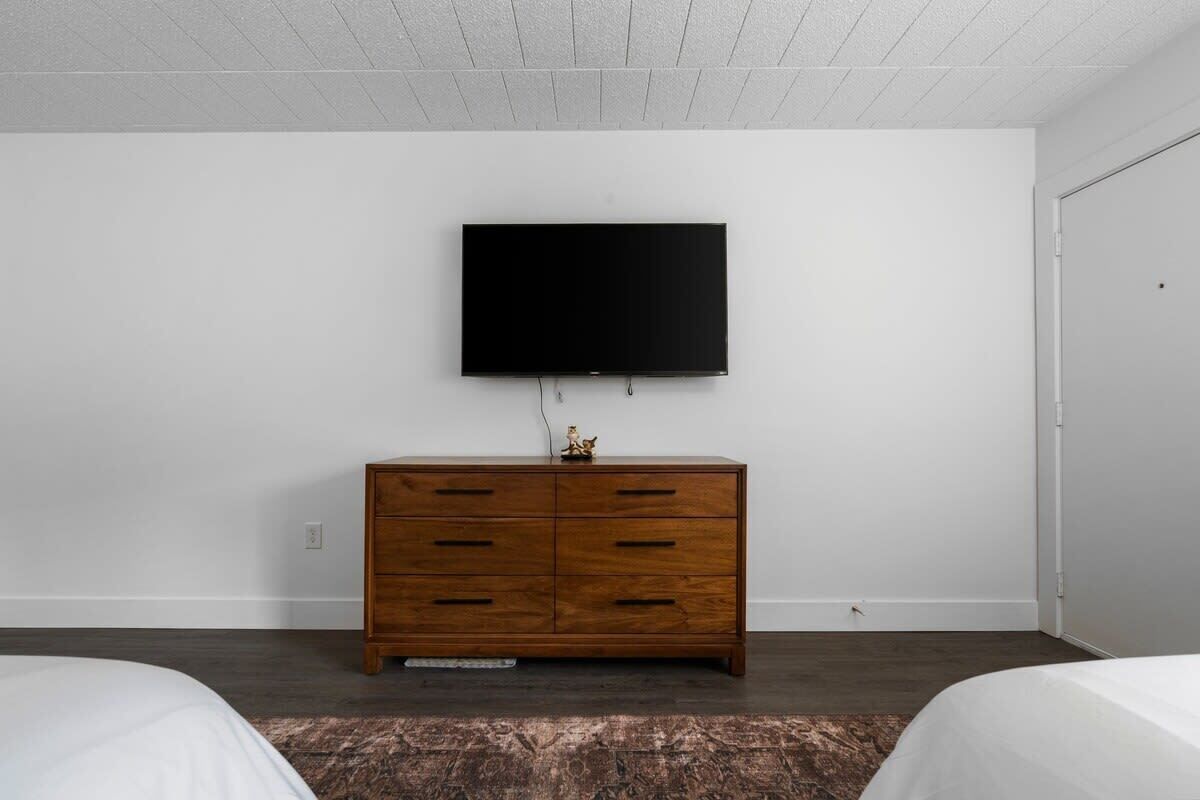 A bedroom with a wooden dresser, a flat-screen TV mounted on the wall, and a bed with a patterned rug.