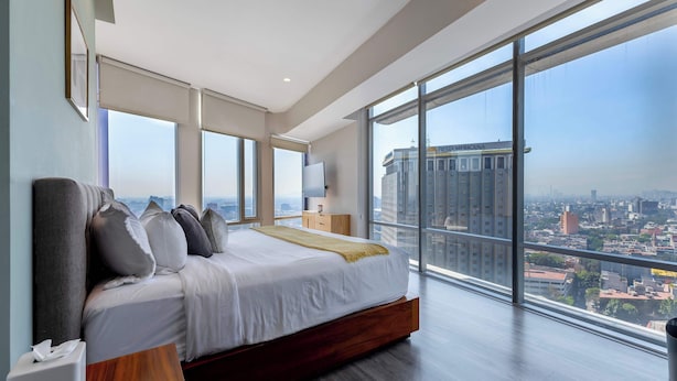 Un dormitorio moderno con una cama grande, una mesita de noche con lámpara y vistas a un paisaje urbano a través de ventanales.