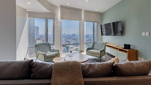 Una sala de estar moderna con un sofá, dos sillones, una mesa de centro y un televisor montado en la pared. Amplias ventanas ofrecen vistas a la ciudad.