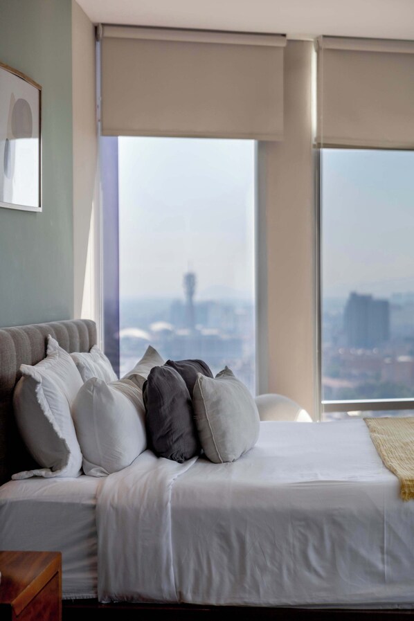Une chambre à coucher avec un lit, des oreillers, une fenêtre donnant sur la ville et un tableau encadré au mur.