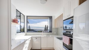 A modern kitchen with white cabinetry, a dishwasher, and a large window offering a view of the outdoors. - Namanula 7 - Oceanstays (Yamba)