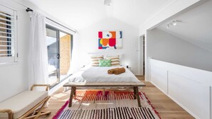 A modern bedroom with a large bed, a ceiling fan, a colorful abstract painting, and a view through a sliding glass door. - Namanula 7 - Oceanstays (Yamba)