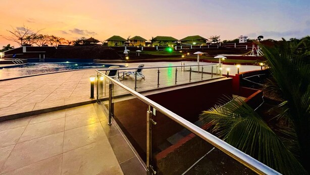 Zona de piscina con vista a un atardecer, sillas de descanso y un edificio con techo verde al fondo.