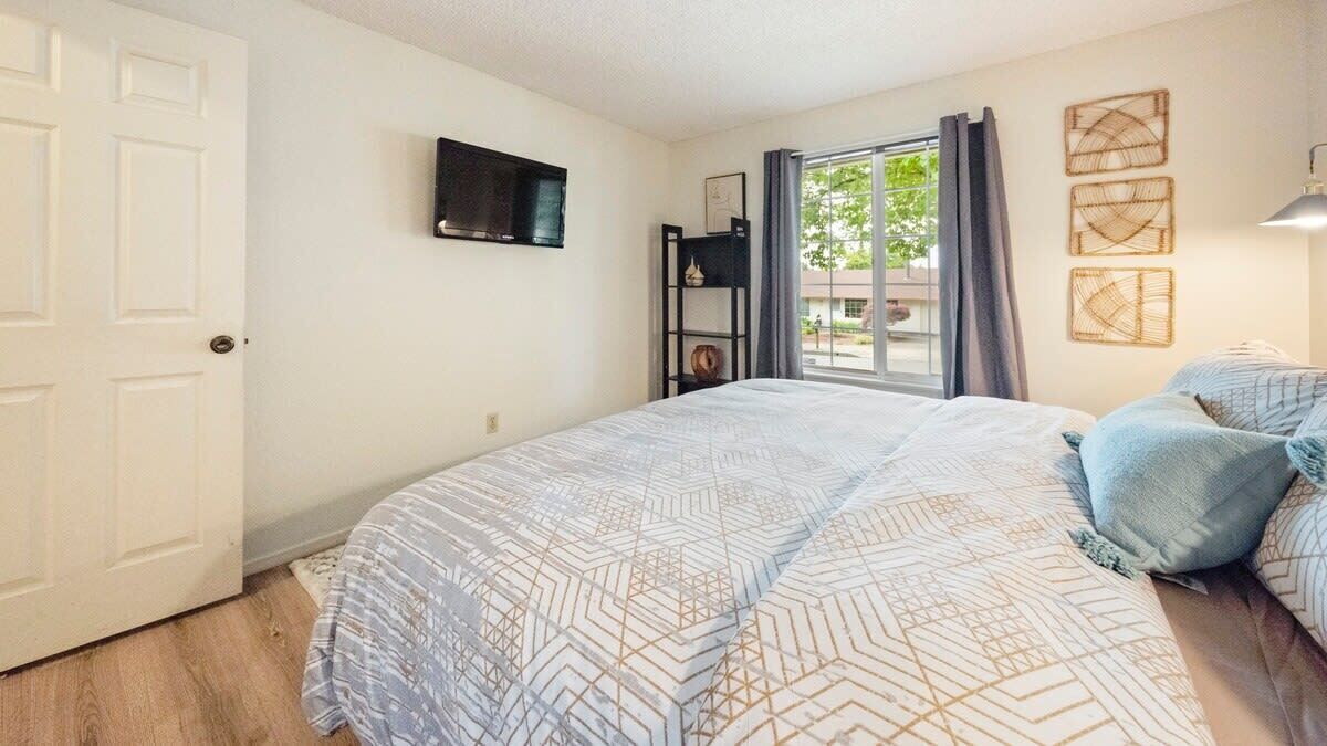 A bedroom with a bed, a TV mounted on the wall, a window with curtains, and a door.