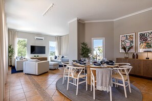 A spacious living area with a dining table set for six, surrounded by white chairs. There is a flat-screen TV mounted on the wall, a sofa set, and a large window offering a view of the outdoors.
