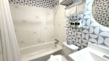 Un baño con bañera, inodoro y lavamanos. Hay una cortina de ducha, un estante de vidrio con artículos de aseo y azulejos estampados en las paredes.