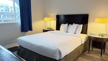 A hotel room with a large bed, two bedside lamps, a window with curtains, and a view of buildings outside.