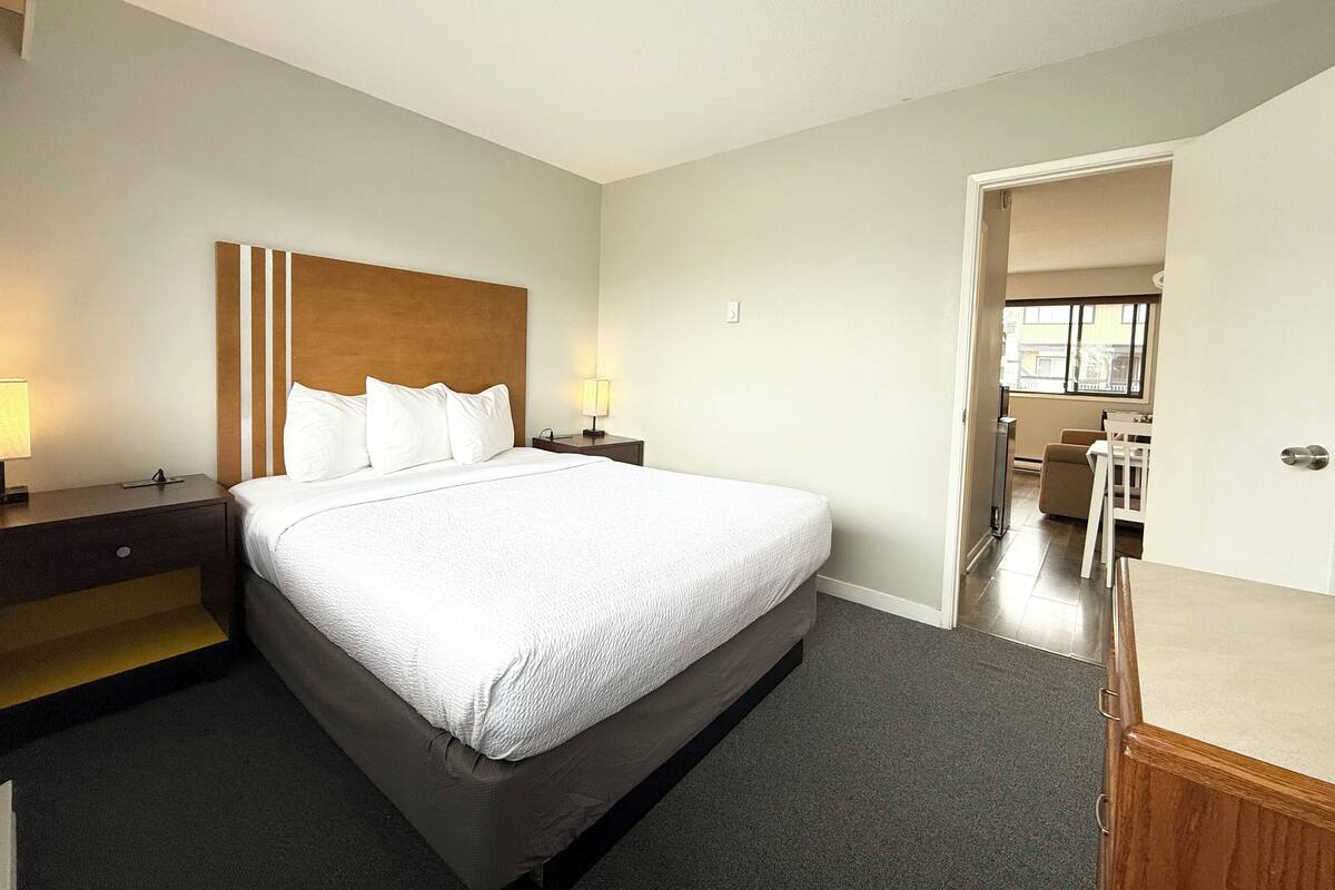 A hotel room with a large bed, a nightstand with a lamp, a wooden headboard, and a view of the bathroom through an open door.