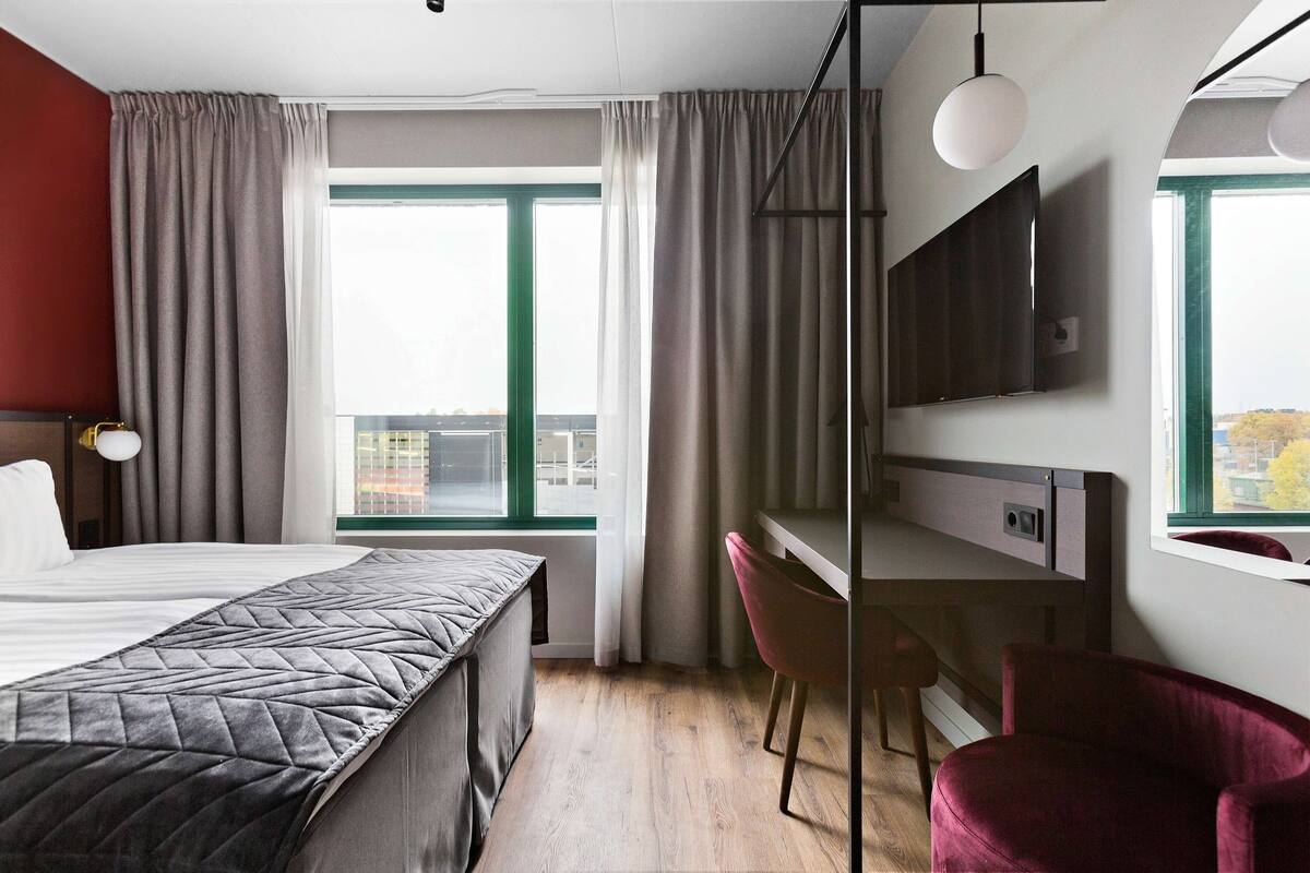 A hotel room with a bed, a desk, a chair, a TV, and a window with curtains.