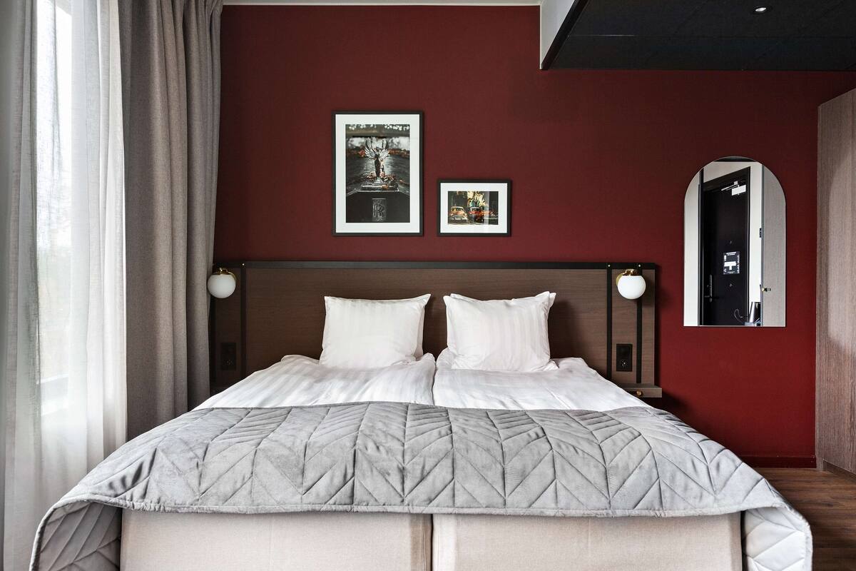 A double bed with a quilted headboard, two framed pictures on the wall, a mirror, and a window with curtains.