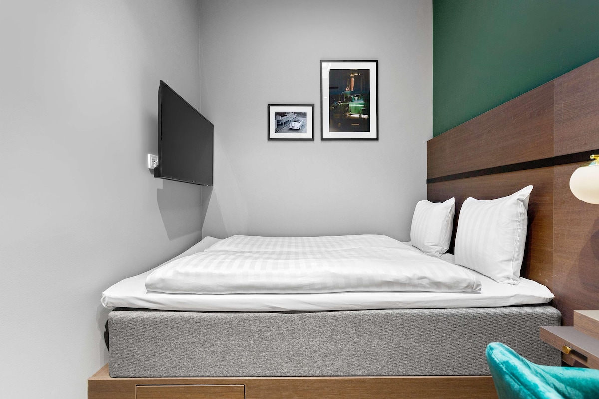 A neatly made bed with white bedding, a grey mattress, and a wooden headboard. There are two framed pictures on the wall, one of a car and another of a building.