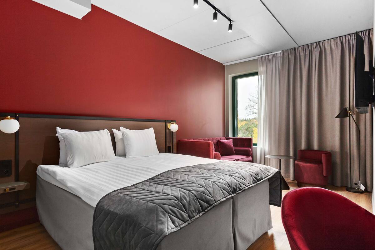 A modern hotel room with a large bed, a red accent wall, and a view of the outdoors through a window.