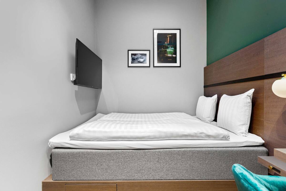 A neatly made bed with white linens, a grey mattress, and a wooden headboard. There are two framed pictures on the wall above the bed.