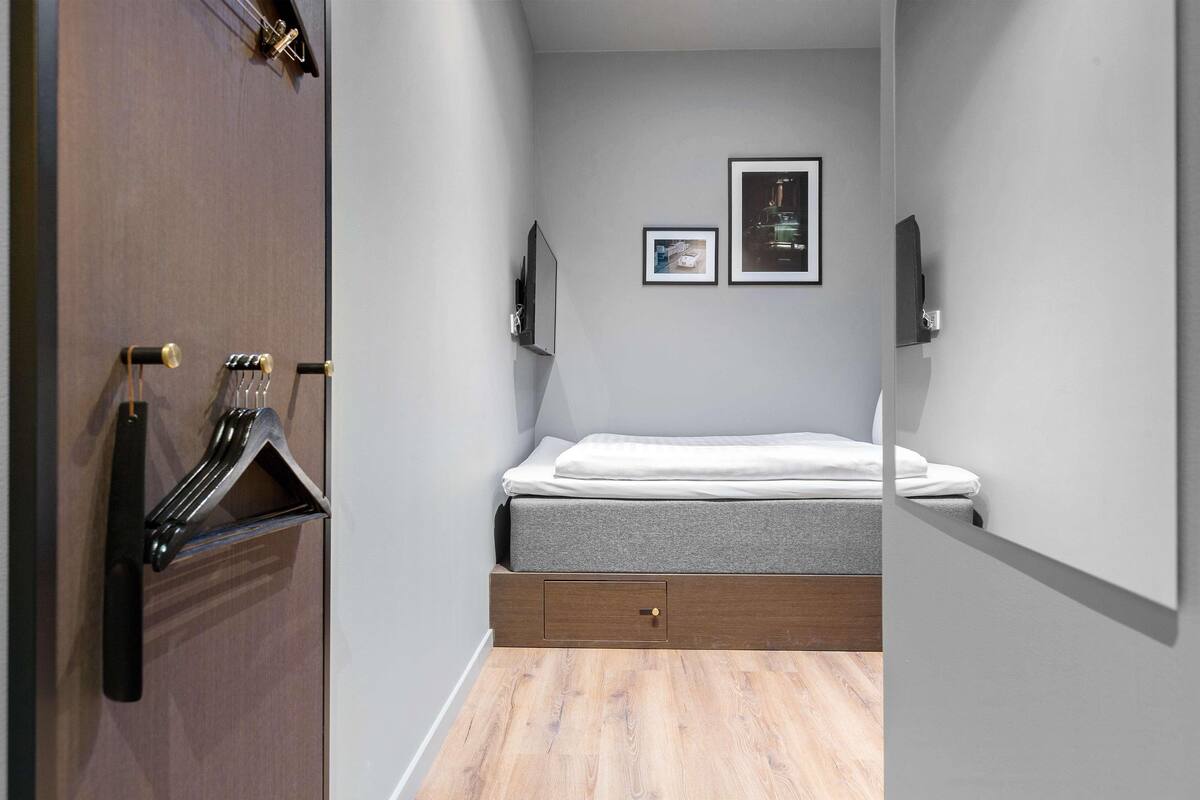 A compact bedroom with a bed, a small bedside table, a wall-mounted TV, and a framed picture.