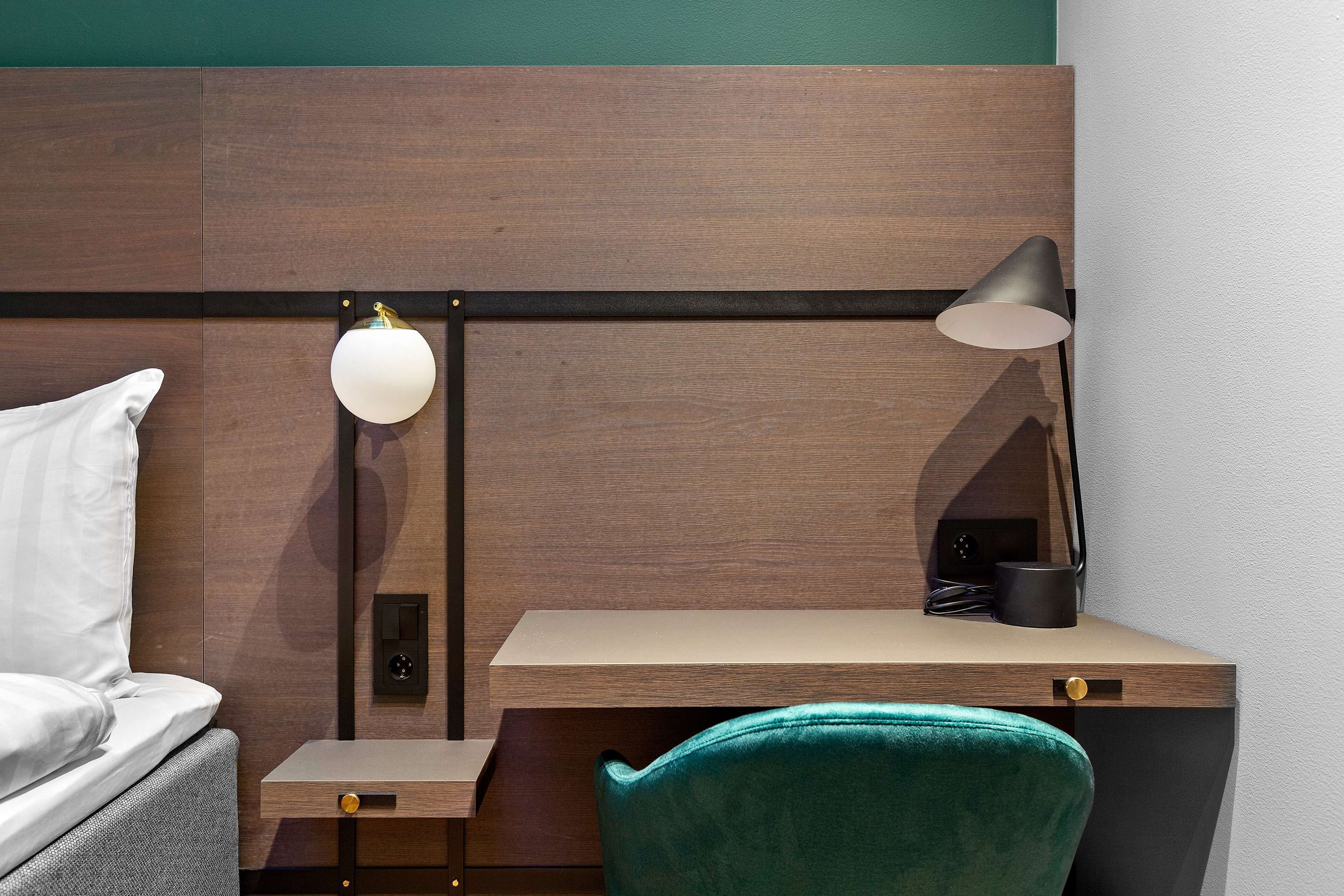 A hotel room with a bed, a desk with a chair, a lamp, and a small table.