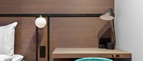 A hotel room with a bed, a desk with a chair, a lamp, and a small table.