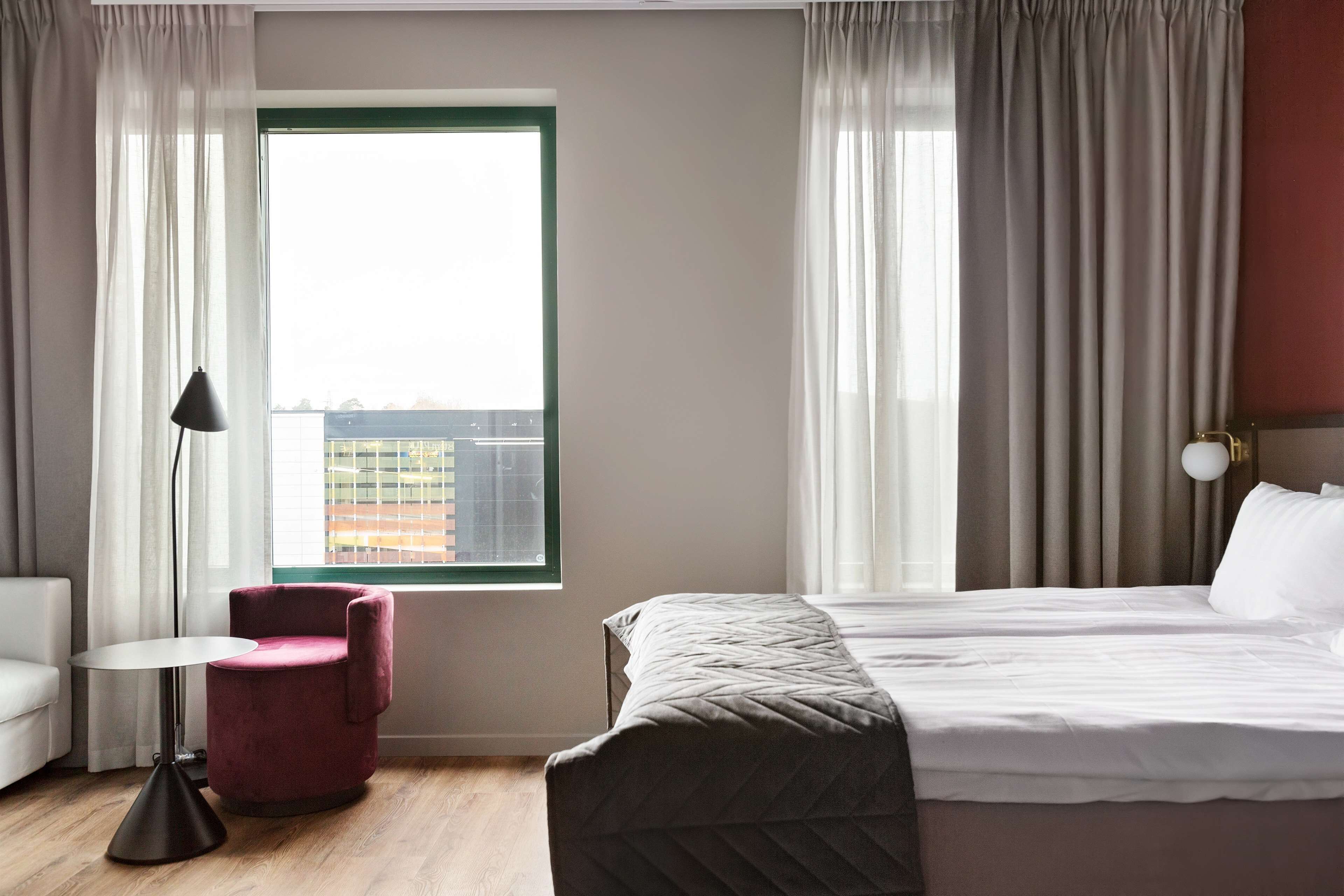 A hotel room with a bed, a chair, a table, and a window with curtains.