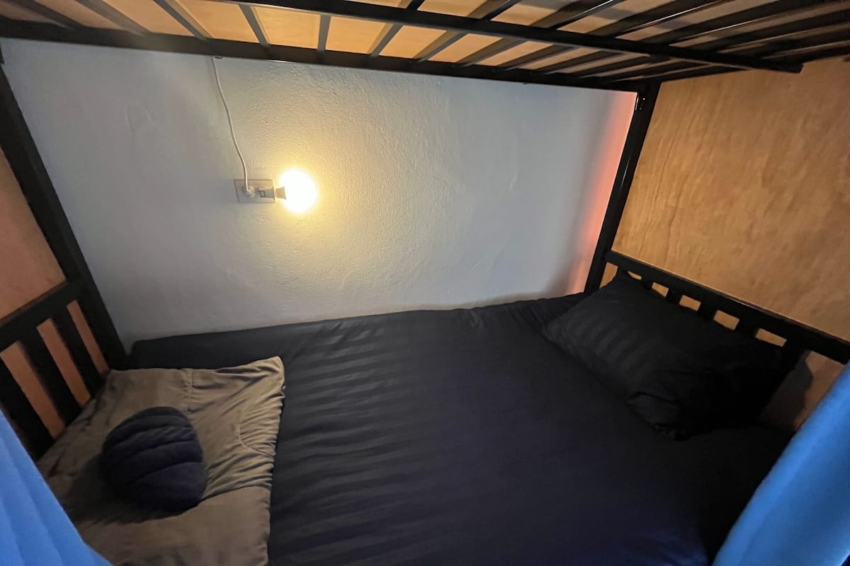 Una habitación pequeña con una cama individual, cabecera de madera, una lámpara en la pared y una cortina a un lado.