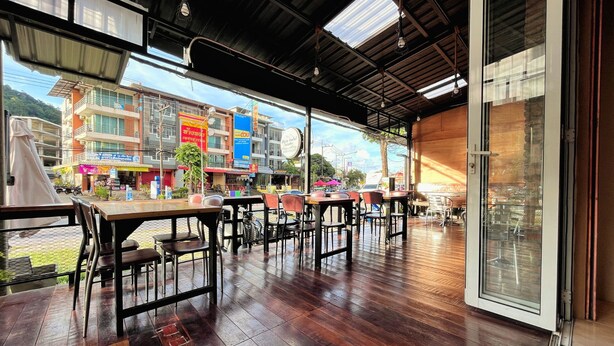 An indoor dining area with wooden tables and chairs, large windows offering a view of the street and buildings outside. - ALPHA Hostel Cafe&Bar (Krabi)