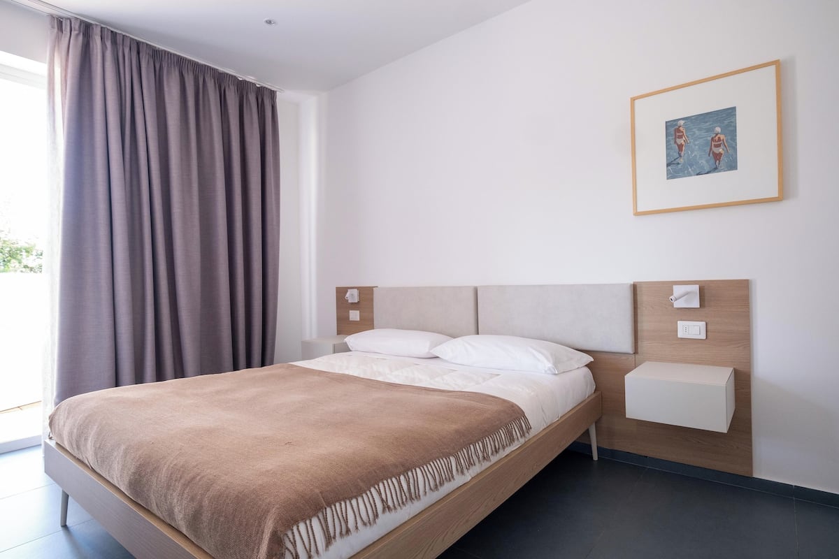 A modern hotel room with a large bed, a bedside table, a wall-mounted light, and a framed picture on the wall.