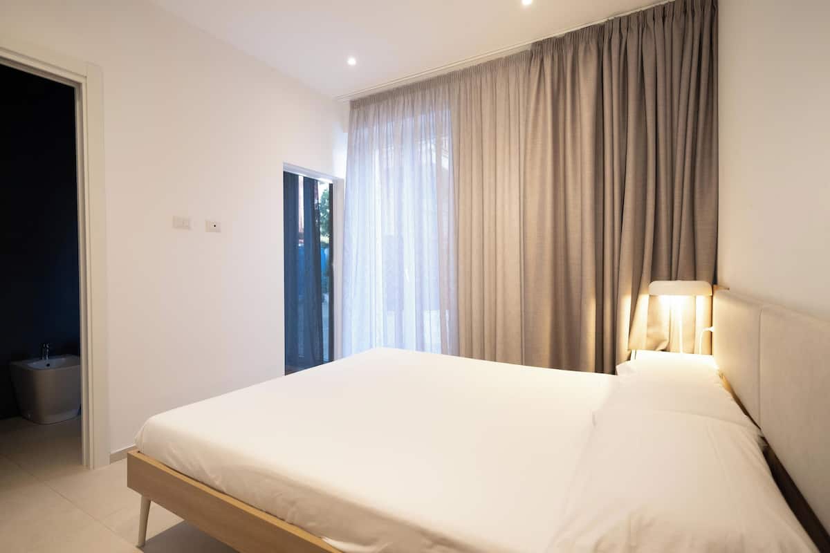 A bedroom with a bed, a window with curtains, a bedside lamp, and a door leading to a bathroom.