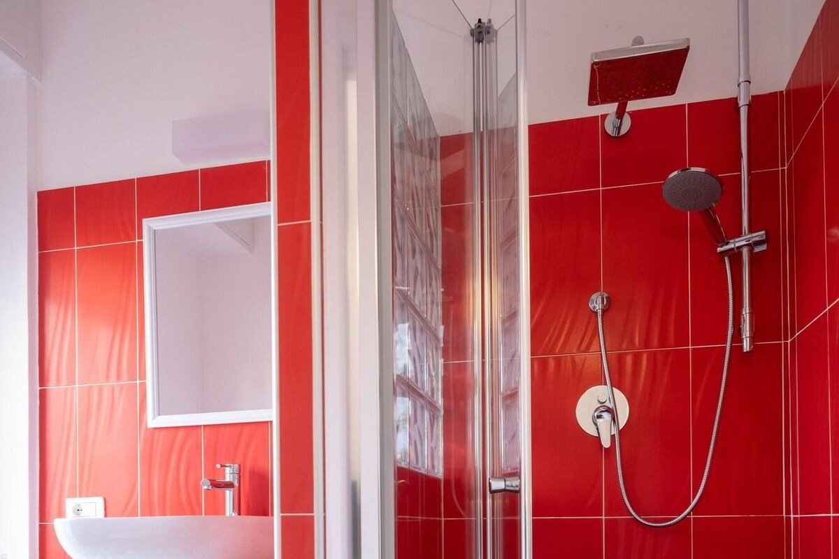 A modern bathroom with a glass-enclosed shower, a white sink, and red tiled walls.