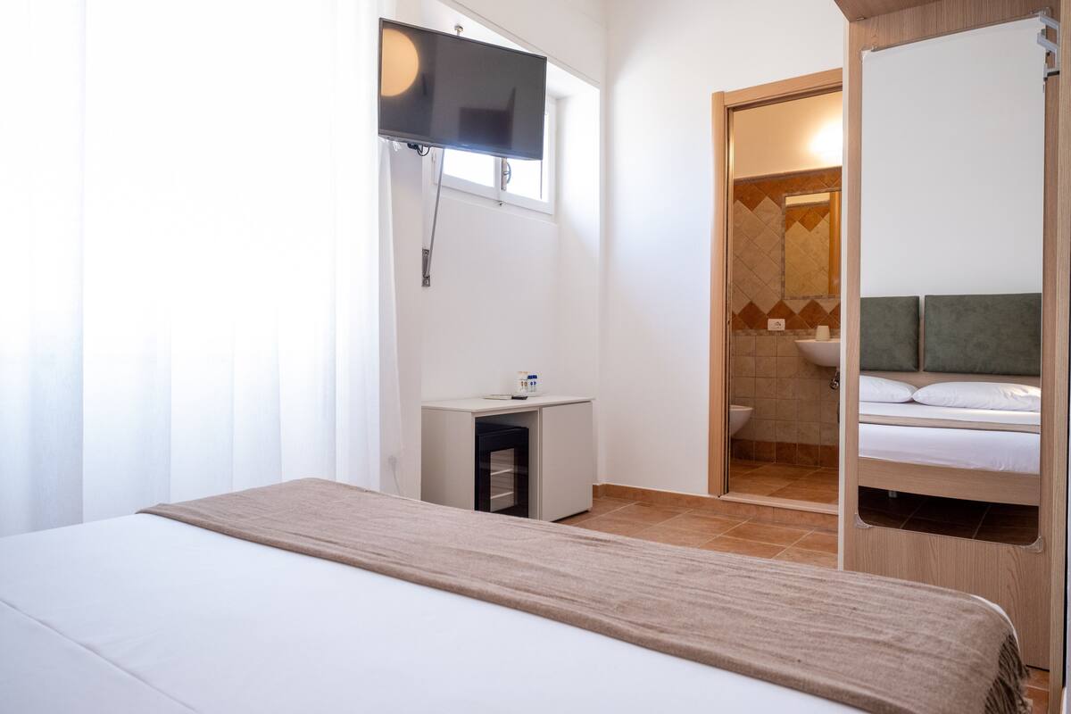 A hotel room with a bed, a TV mounted on the wall, a small desk, and a bathroom visible through an open door.
