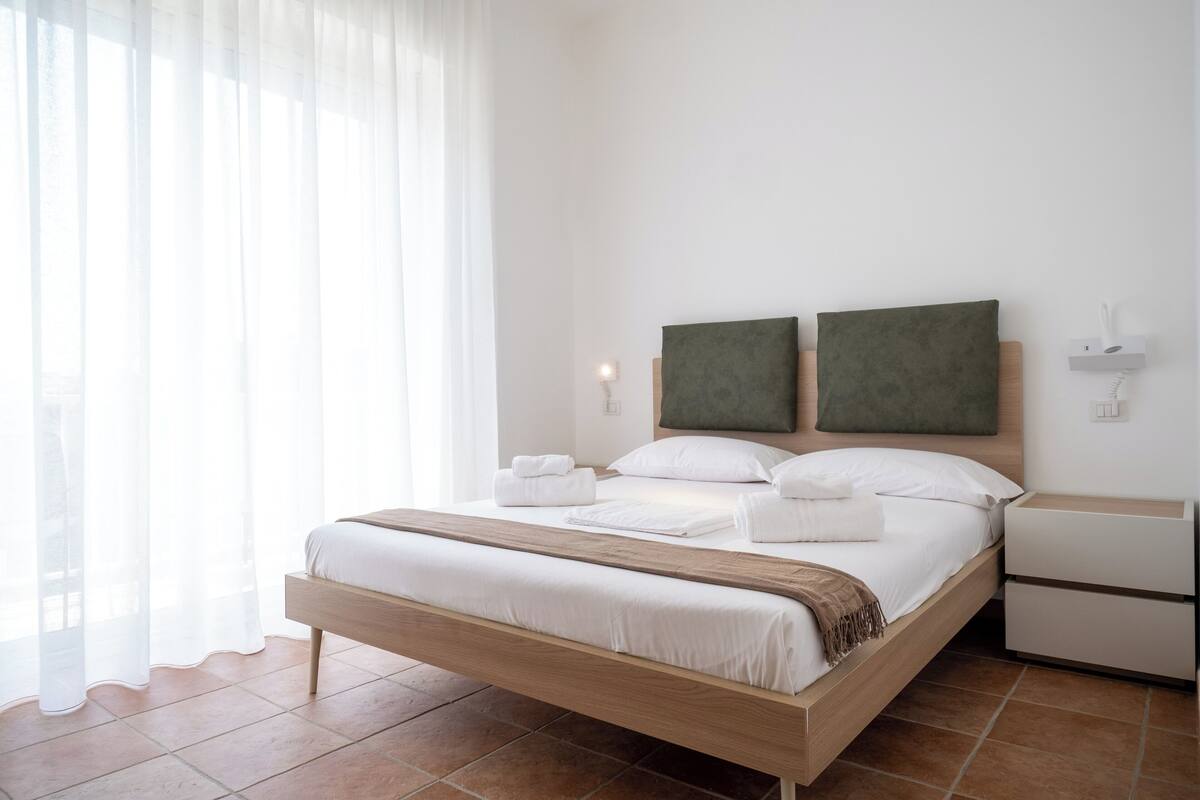A modern hotel room with a wooden bed, white linens, a large window with sheer curtains, and a minimalist decor.