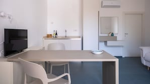 A modern, minimalist room with a flat-screen TV, a dining table, a white chair, a small kitchen area with a sink and a mirror, and a door. - Nautika Suites  (Alghero)