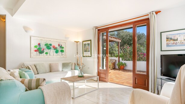 Un salon spacieux comprenant un canapé, une table basse et un téléviseur. Une porte donne accès à un espace extérieur agrémenté de plantes et offrant une vue sur la mer.