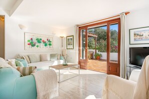 Un salon spacieux comprenant un canapé, une table basse et un téléviseur. Une porte donne accès à un espace extérieur agrémenté de plantes et offrant une vue sur la mer.