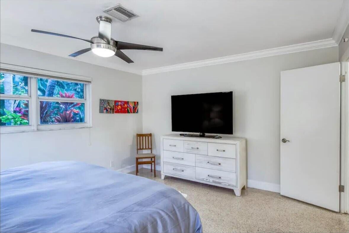A bedroom with a bed, a TV on a dresser, a ceiling fan, and a window with a view of greenery.