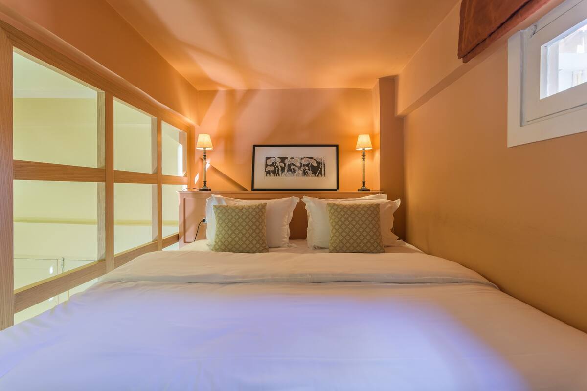 A neatly made bed with white linens and two green patterned pillows, a framed artwork above the bed, and two bedside lamps.