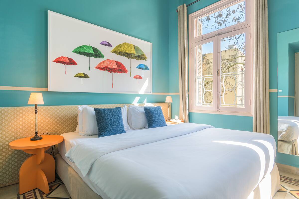 A bed with white bedding, a blue and white patterned headboard, and a painting of colorful umbrellas on the wall.