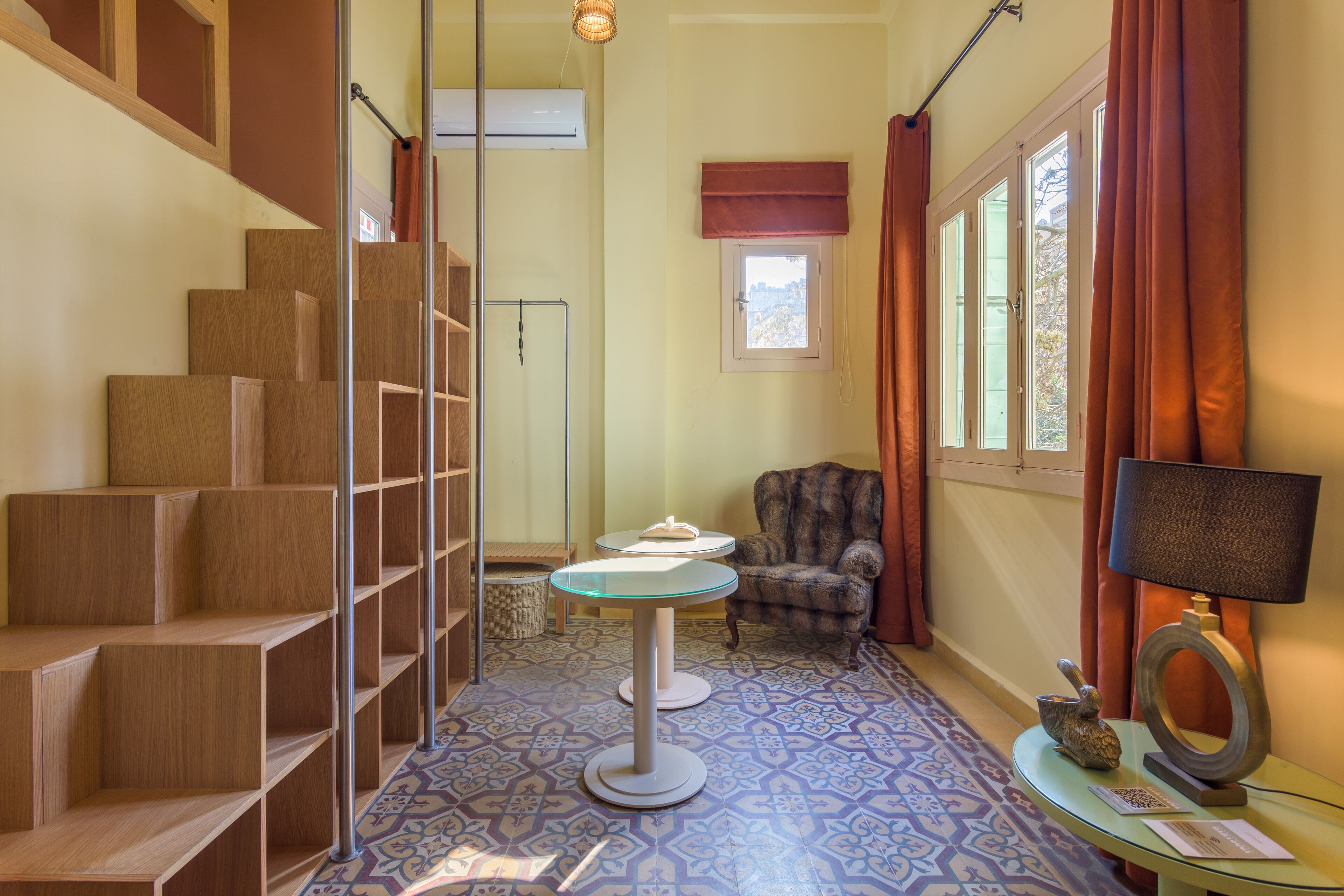 A room with a wooden staircase, a patterned floor, a small table with a lamp, and a chair.